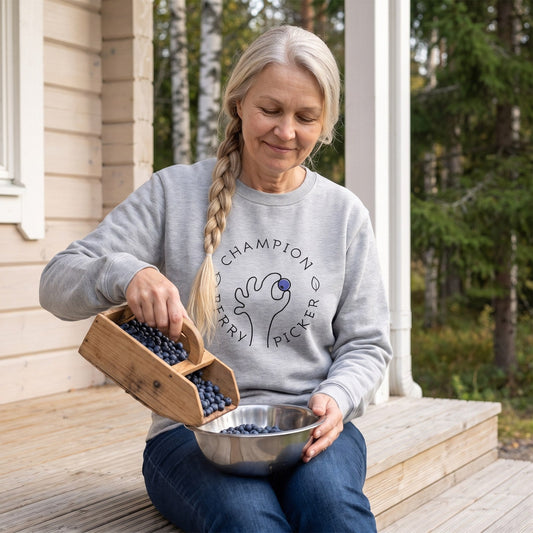 Champion Blueberry Picker sweatshirt in sport grey, worn by Anneli tipping blueberries into a bowl on a cottage veranda, Very Finnish Problems