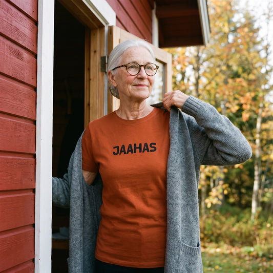 Jaahas Finnish language t-shirt in burnt orange, worn by a woman pulling on a cardigan in the doorway of a red summer cottage, Very Finnish Problems