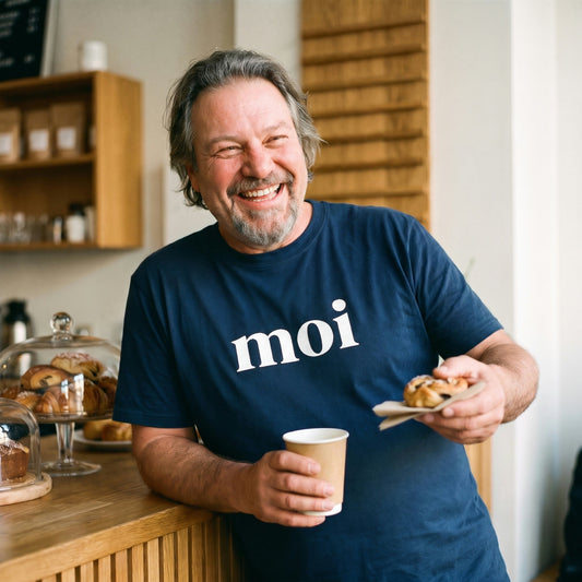 Moi Finnish greeting t-shirt in navy, worn by Jojelli laughing at a Finnish cafe counter with a coffee and a pastry, Very Finnish Problems
