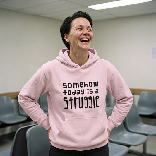 Somehow today is a struggle Finnish culture hoodie in light pink, worn by Katja laughing in a waiting room, Very Finnish Problems