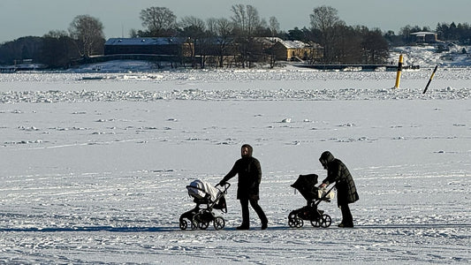Two people pushing prams across a frozen sea in bright winter sunshine near Helsinki, with a snow covered shoreline behind them