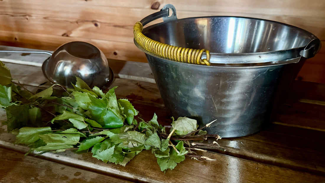 Steel sauna bucket with birch whisk on a wooden bench inside a Finnish sauna, photo by Joel Willans