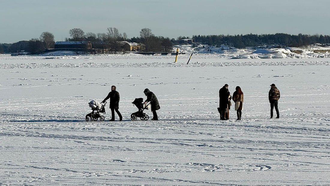 Two parents pushing prams across the frozen sea in Helsinki on a bright winter day, with small groups of people spaced out on the ice and the snowy shoreline behind