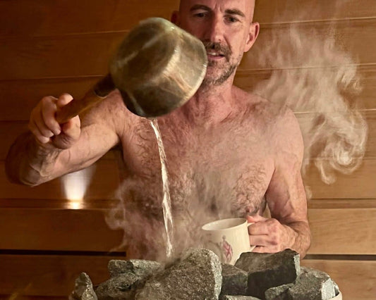 A man throwing a ladle of water onto the hot stones of a Finnish sauna, steam rising off the kiuas. Photo: Joel Willans