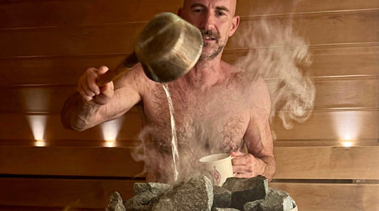 A man throwing a ladle of water onto the hot stones of a Finnish sauna, steam rising off the kiuas