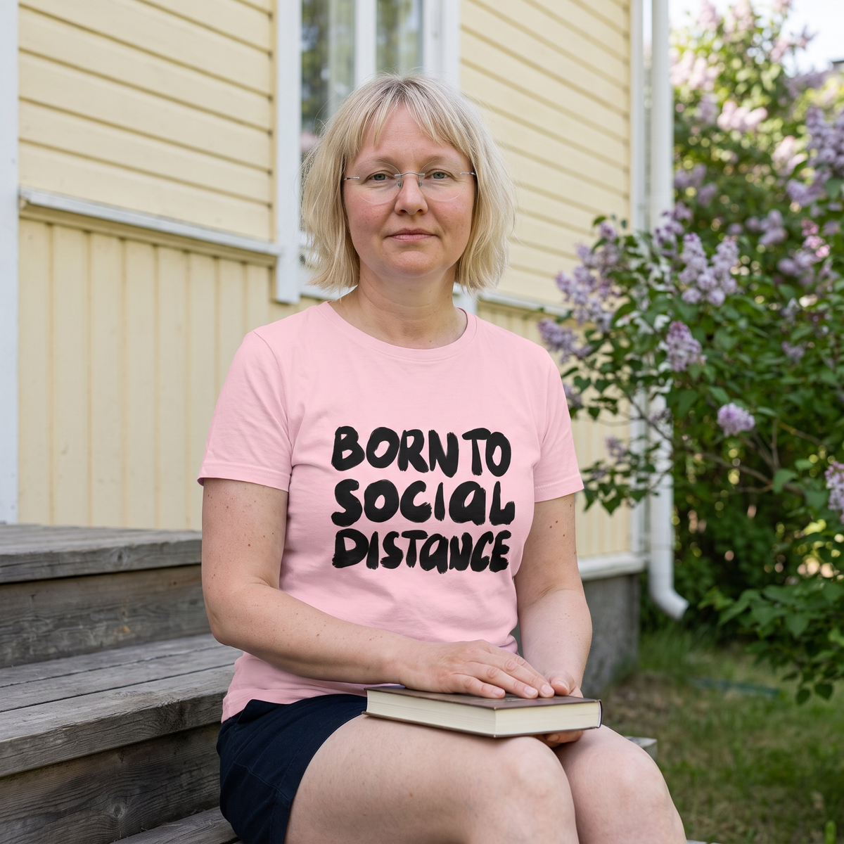 Born to Social Distance t-shirt in pink, worn by Marika on the porch steps of a wooden house with a book, Very Finnish Problems