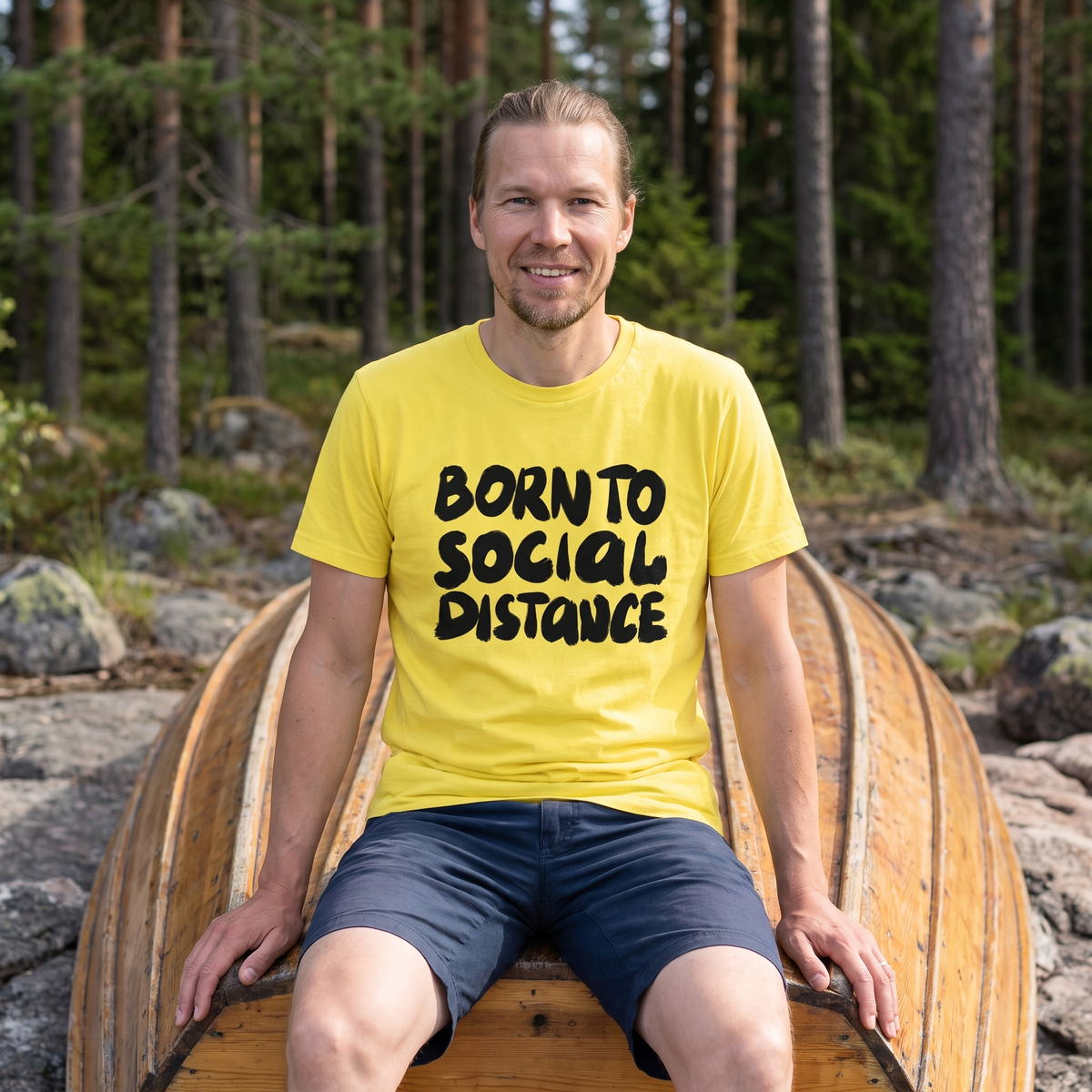 Born to Social Distance t-shirt in yellow, worn by Joonas sitting on an upturned rowing boat on a deserted lakeshore, Very Finnish Problems