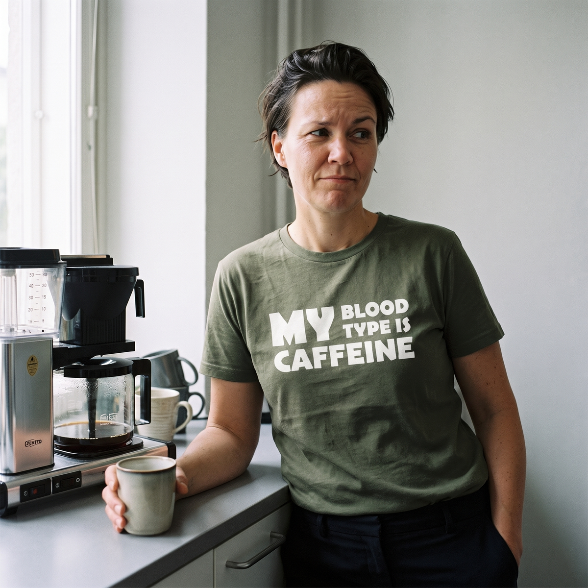 Caffeine Blood Type t-shirt in green olive, worn by Katja leaning on an office kitchenette counter at the afternoon coffee break, Very Finnish Problems