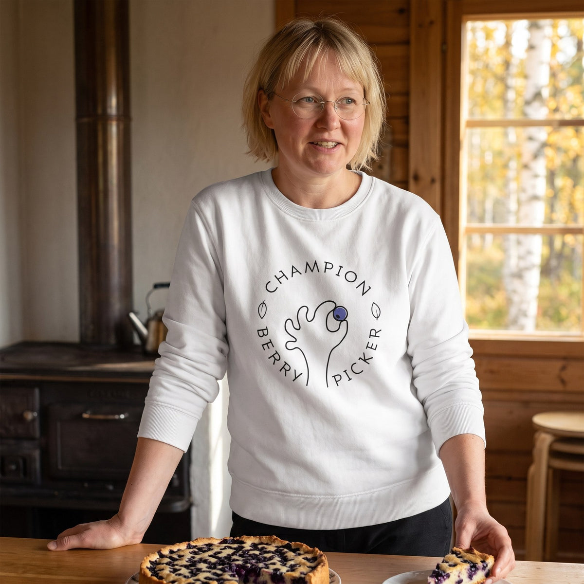 Champion Blueberry Picker sweatshirt in white, worn by Marika beside a blueberry pie in a cottage kitchen, Very Finnish Problems