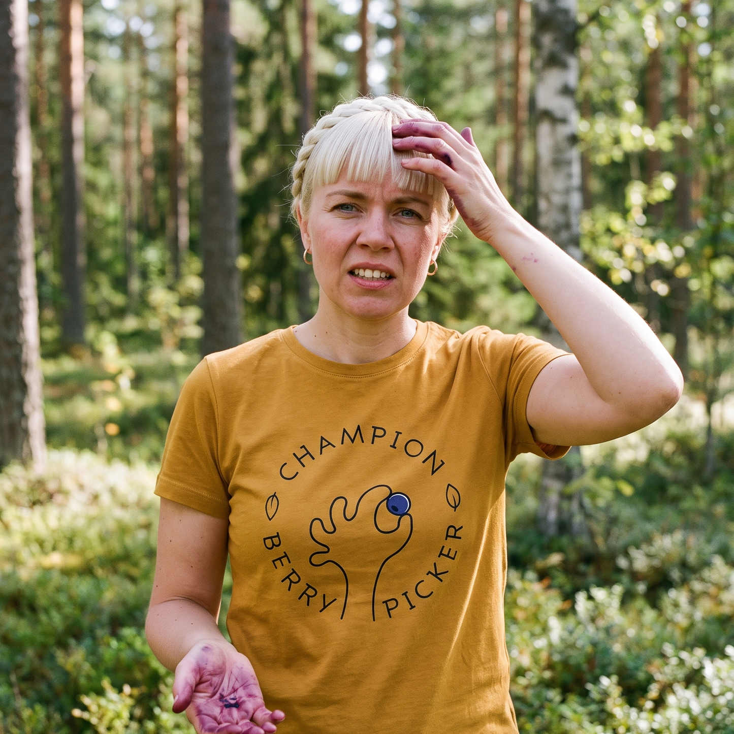 Champion Blueberry Picker t-shirt in gold, worn by Venla with berry-stained fingers in a forest clearing, Very Finnish Problems