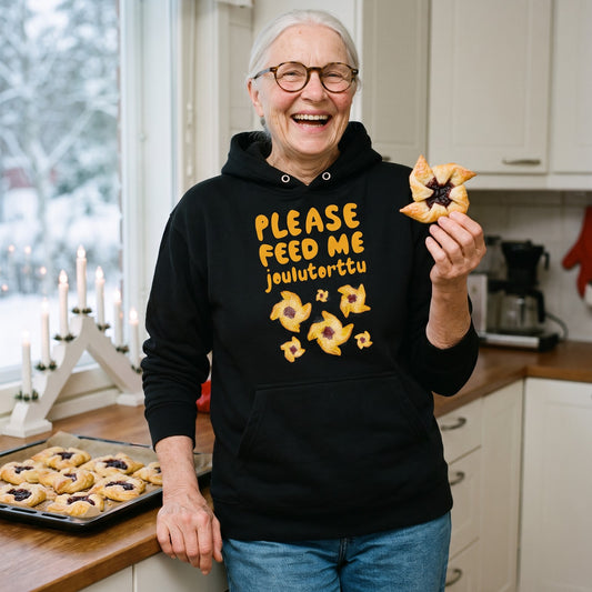 Feed me Joulutorttu Finnish Holiday Hoodie in black, worn by Raija laughing with a joulutorttu in a Christmas kitchen, Very Finnish Problems