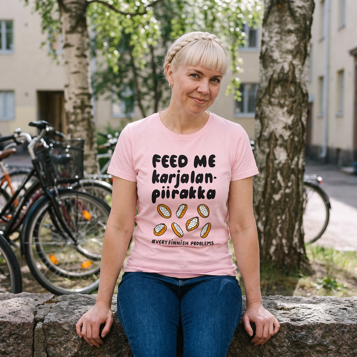 Feed Me Karjalanpiirakka t-shirt in pink, worn by Venla sitting on a stone wall in a Helsinki courtyard, Very Finnish Problems