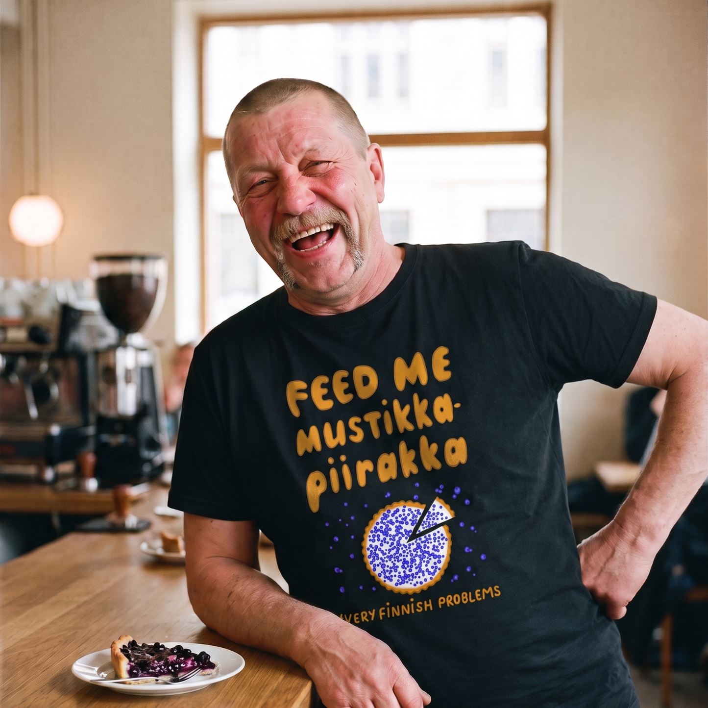Feed Me Mustikkapiirakka t-shirt in black, worn by Juha laughing at a cafe counter with a slice of blueberry pie, Very Finnish Problems
