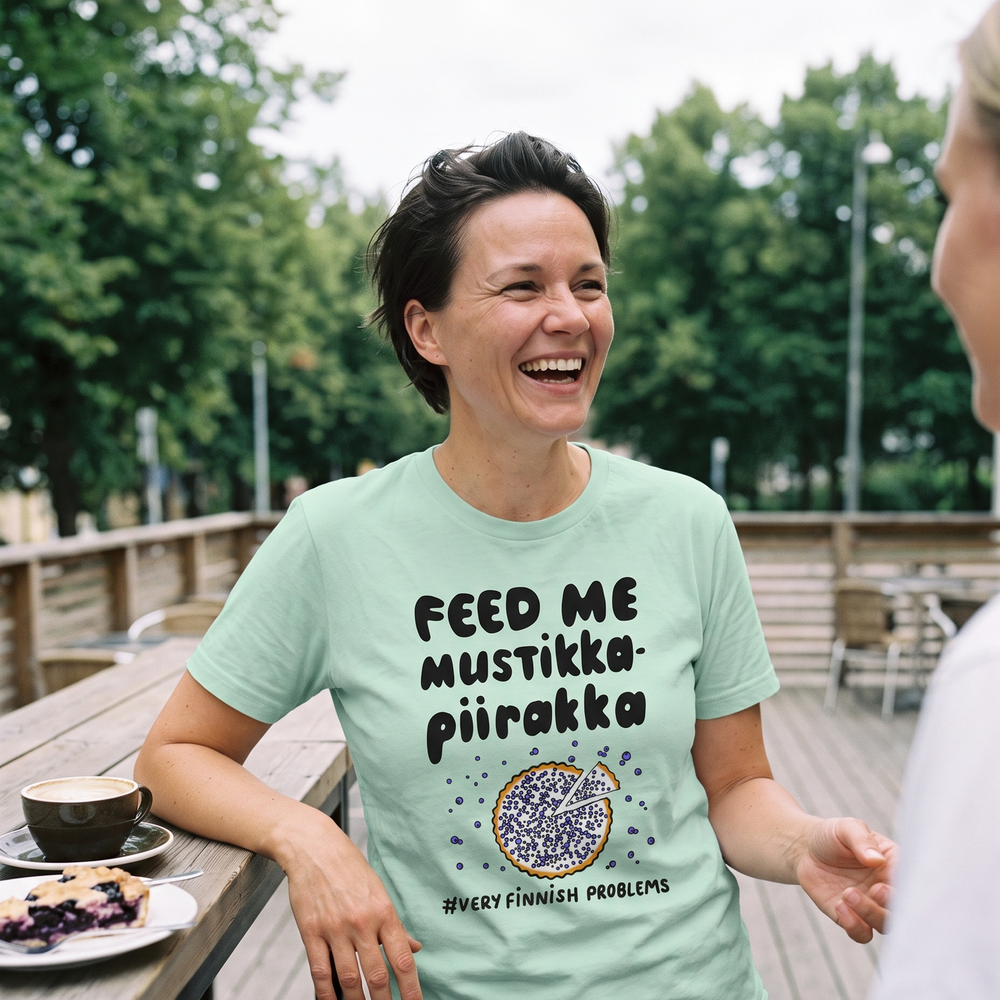 Feed Me Mustikkapiirakka t-shirt in mint, worn by Katja on a summer cafe terrace with coffee and pie, Very Finnish Problems