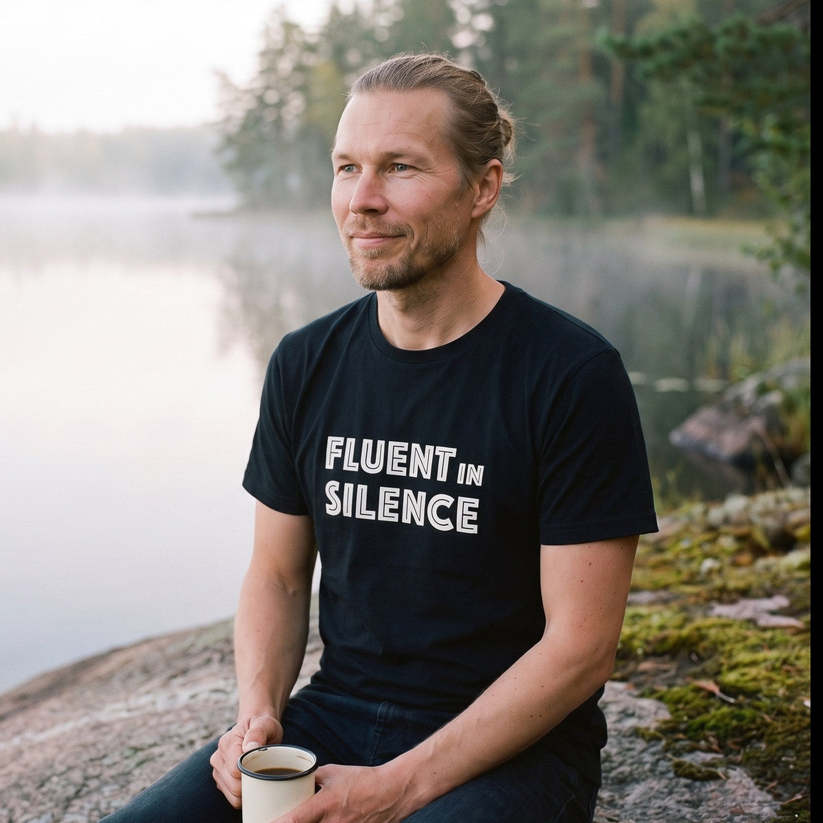 Fluent in Silence t-shirt in black, worn by Joonas with a coffee on a misty lakeside rock at dawn, Very Finnish Problems