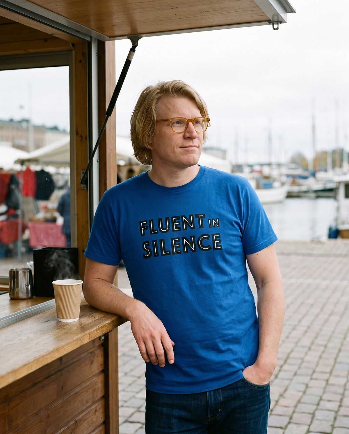 Fluent in Silence t-shirt in true royal, worn by Tuomas leaning on a market square coffee kiosk, Very Finnish Problems