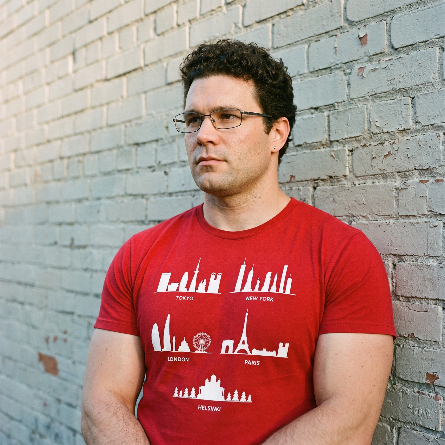 Helsinki Skyline t-shirt in red, worn by Tuomas against a painted brick wall, Very Finnish Problems