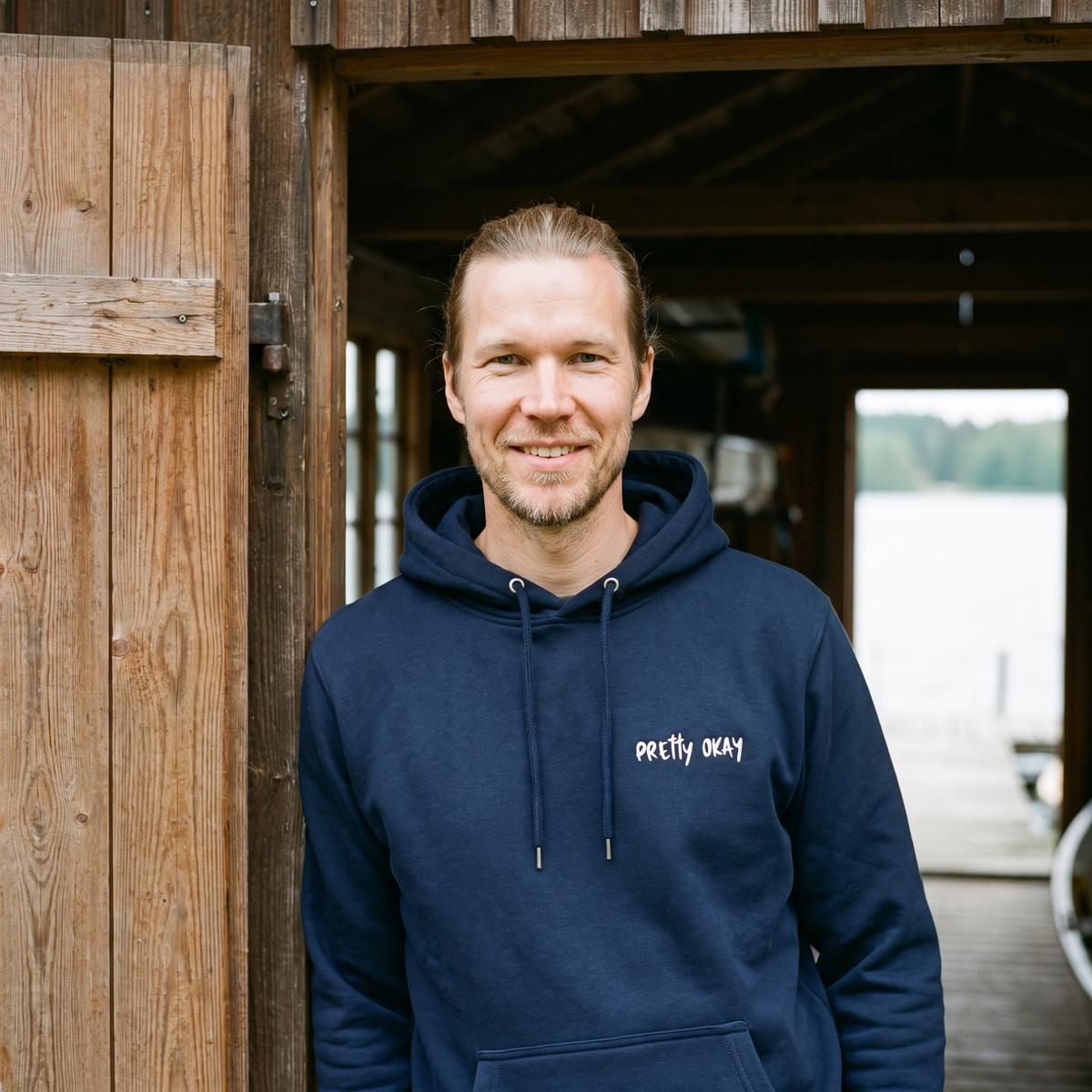 Pretty okay embroidered Finnish humour hoodie in navy, worn by Joonas in a boat shed doorway, Very Finnish Problems