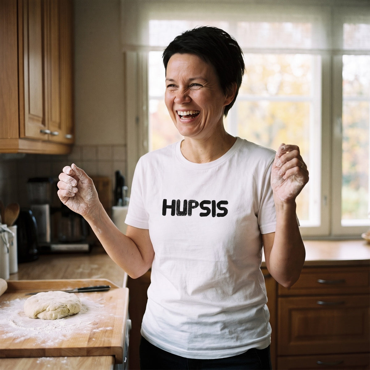 Hupsis Finnish language t-shirt in white, worn by a woman with floury hands laughing at herself in a kitchen, Very Finnish Problems