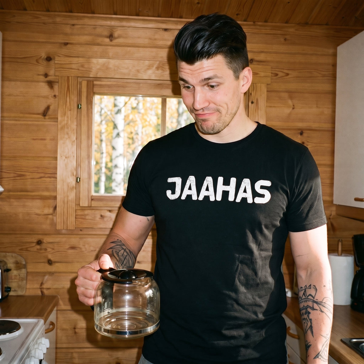 Jaahas Finnish language t-shirt in black, worn by a man raising his eyebrows at an empty coffee pot in a cottage kitchen, Very Finnish Problems