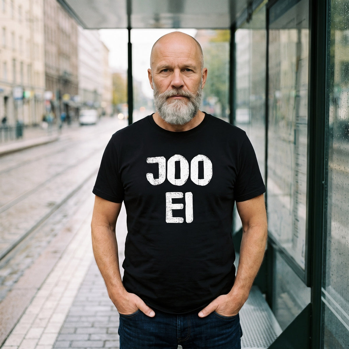 Joo ei Finnish language t-shirt in black, worn by a deadpan man with his thumbs in his pockets at a rainy tram stop, Very Finnish Problems