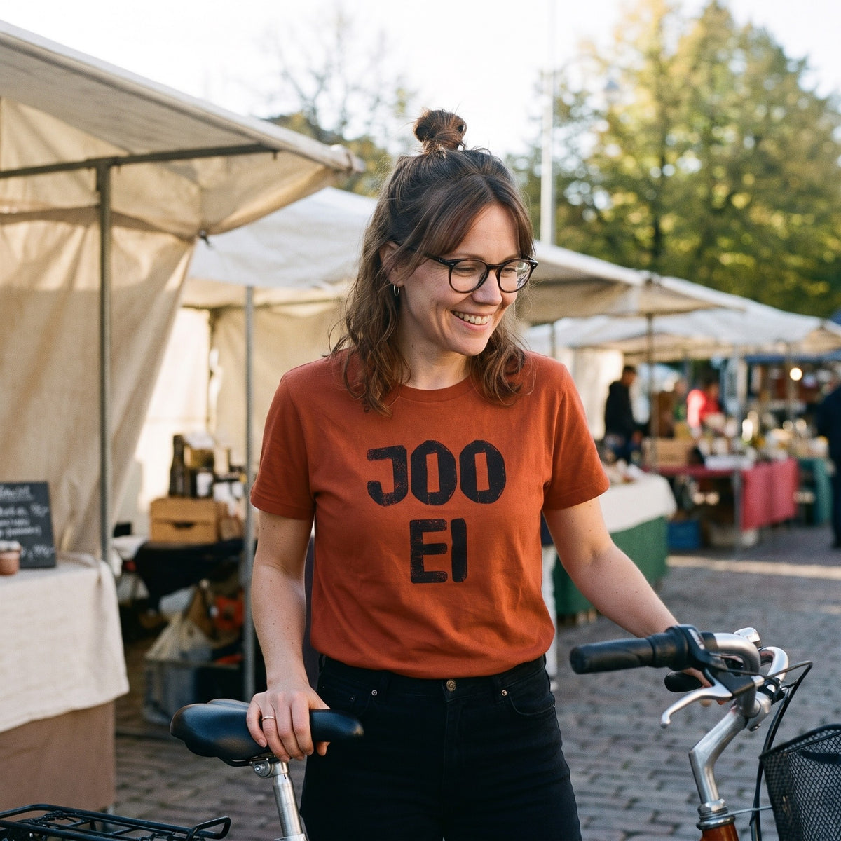 Joo ei Finnish language t-shirt in burnt orange, worn by a woman shaking her head with a smile beside her bike at a market stall, Very Finnish Problems