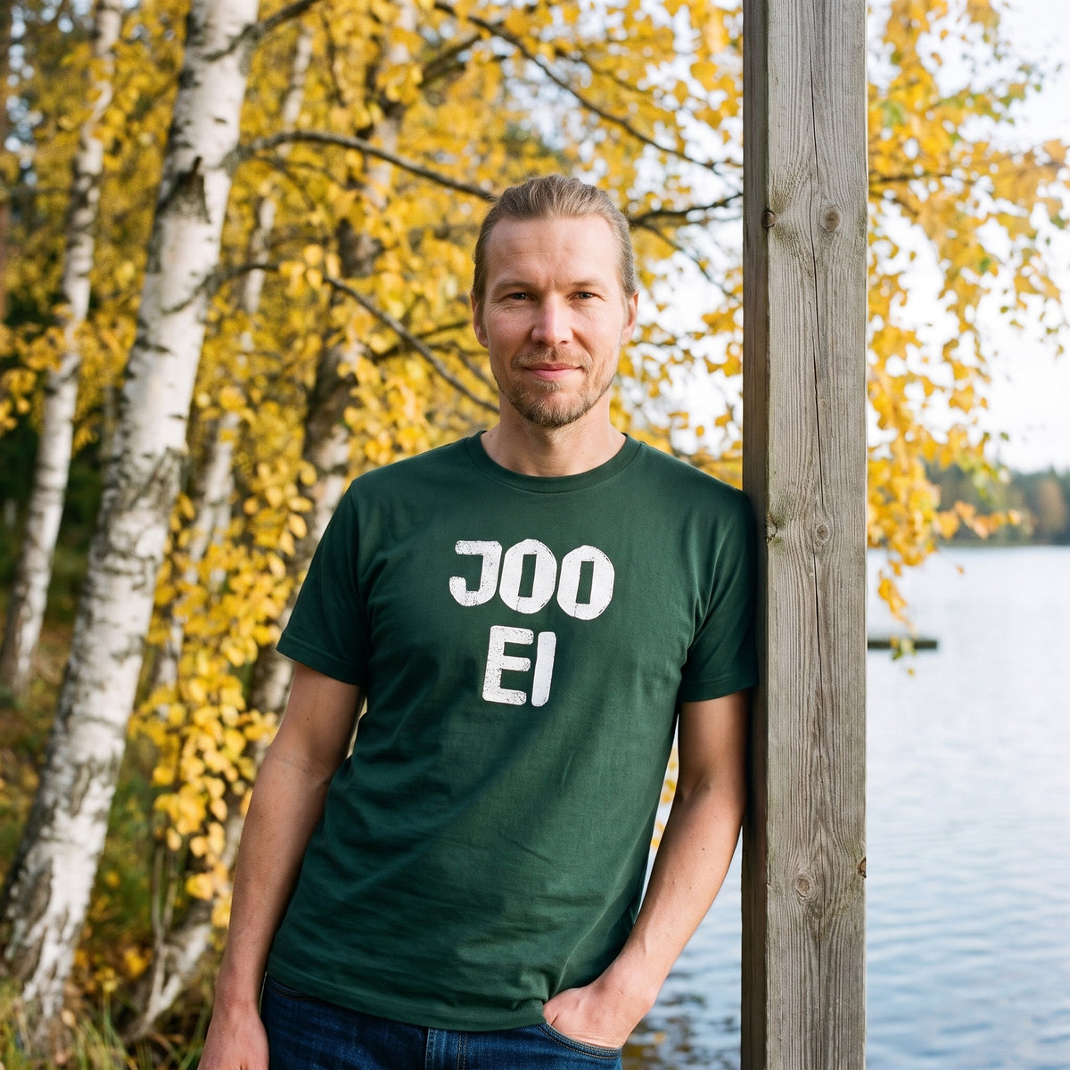 Joo ei Finnish language t-shirt in forest green, worn by a man leaning on a jetty post by autumn birches, Very Finnish Problems