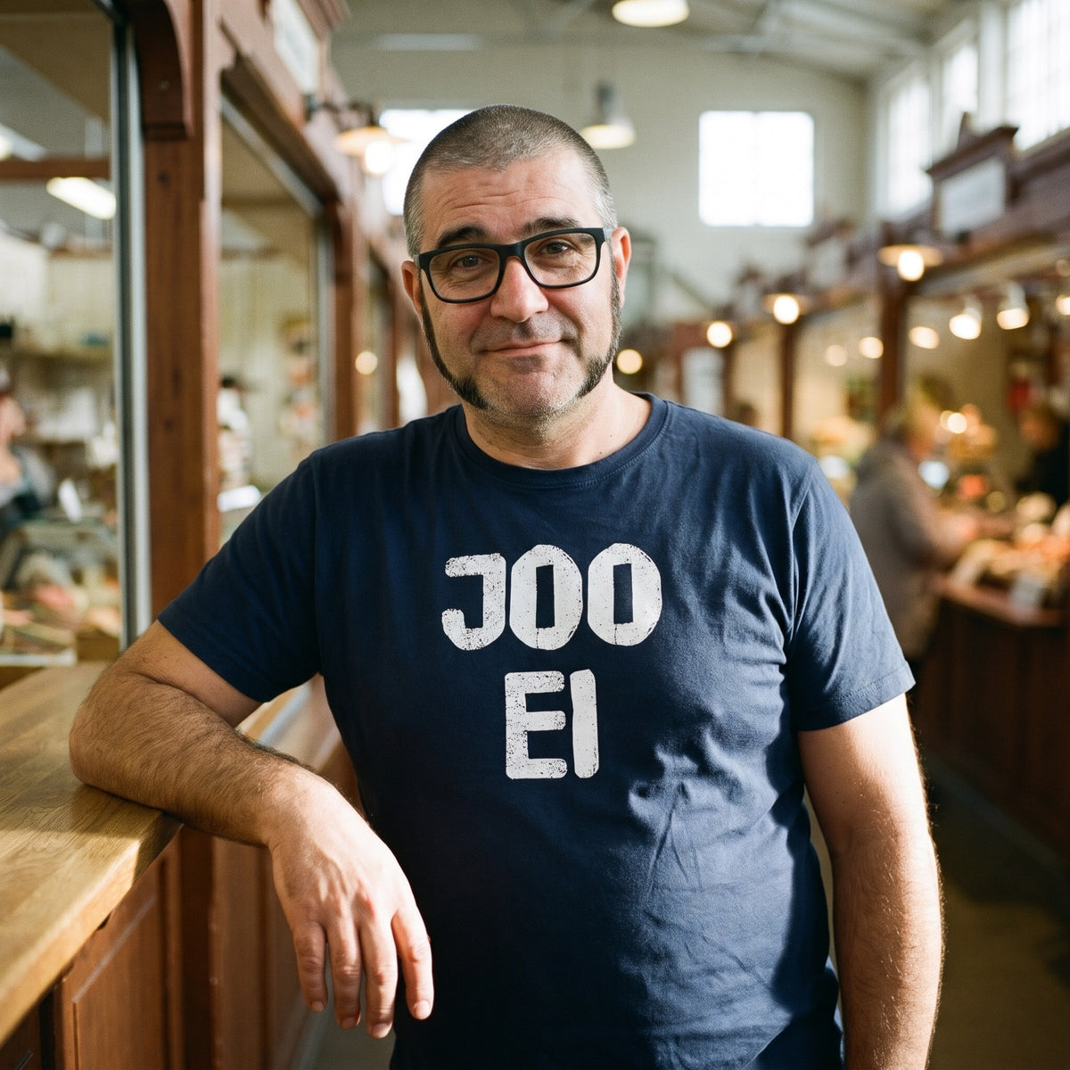 Joo ei Finnish language t-shirt in navy, worn by a man with a wry raised eyebrow leaning on a market hall counter, Very Finnish Problems