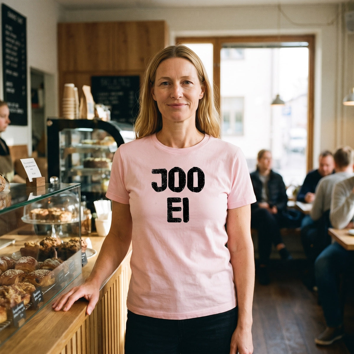 Joo ei Finnish language t-shirt in pink, worn by a woman giving a polite but firm no at a café counter, Very Finnish Problems