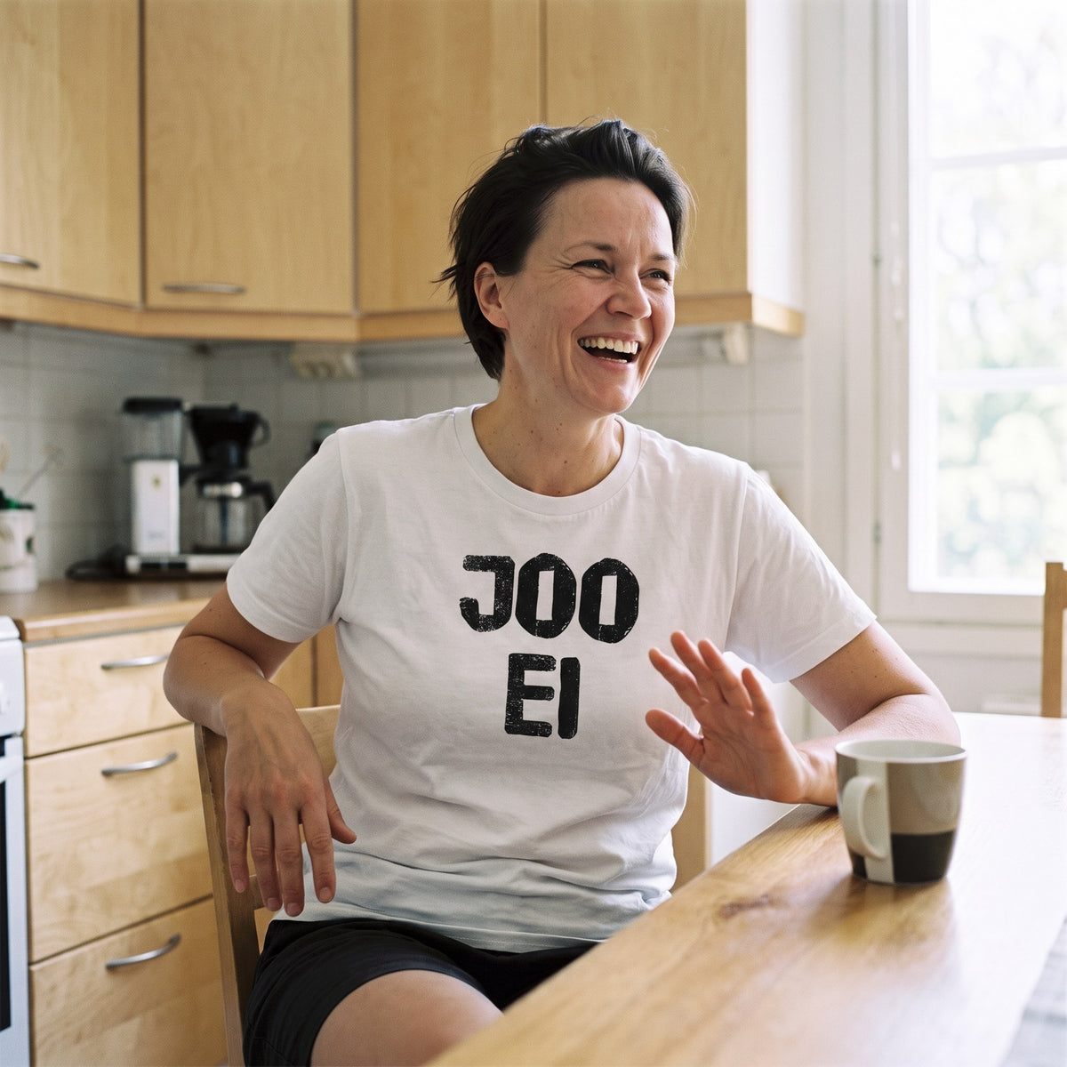 Joo ei Finnish language t-shirt in white, worn by a woman raising a hand in a polite no at a kitchen table, Very Finnish Problems