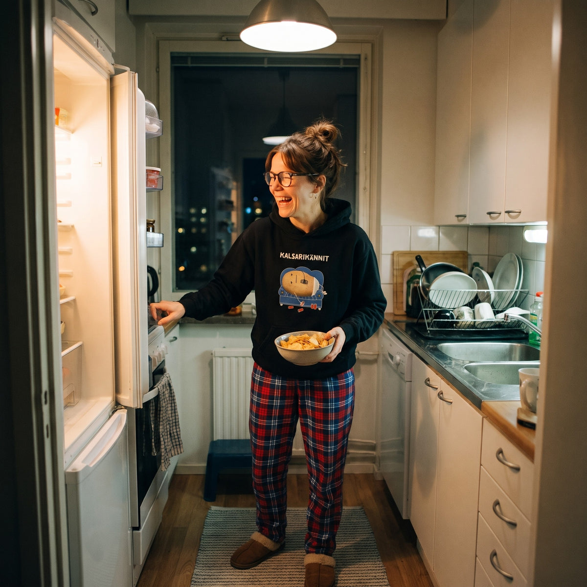Kalsarikännit hoodie in black, worn by a laughing woman in pyjama trousers at an open fridge with a bowl of crisps, Very Finnish Problems