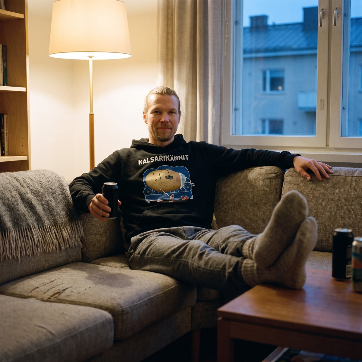 Kalsarikännit hoodie in black, worn by a man with a man-bun sunk into a sofa with his feet on the coffee table, Very Finnish Problems
