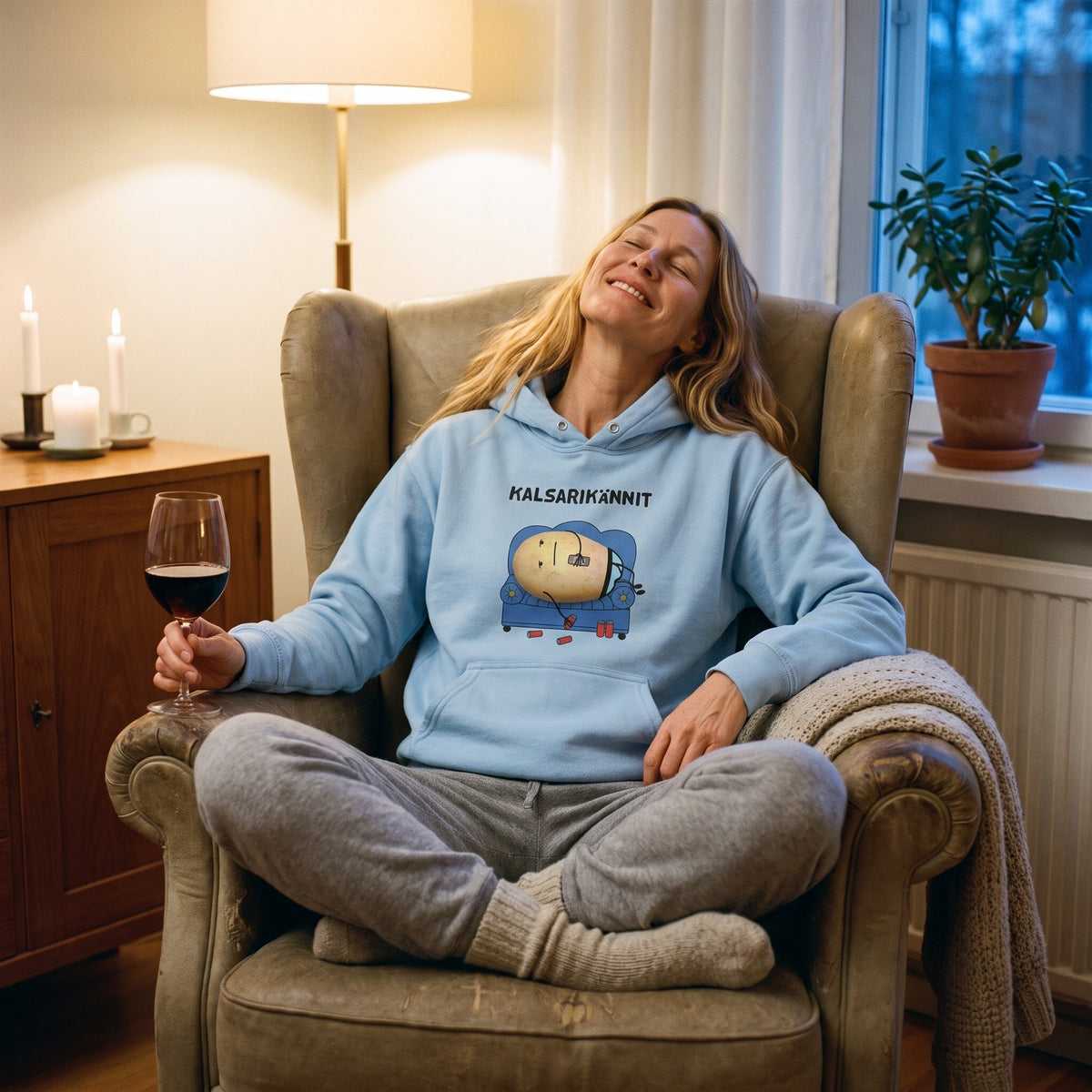Kalsarikännit hoodie in light blue, worn by a woman curled up in an armchair with a glass of red wine, Very Finnish Problems