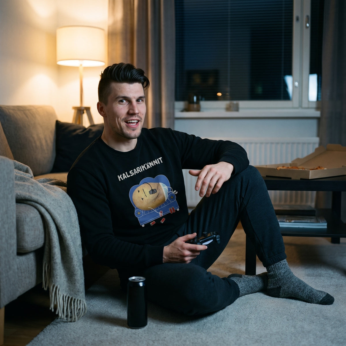 Kalsarikännit sweatshirt in black, worn by a grinning man on the living room floor with a games controller, Very Finnish Problems