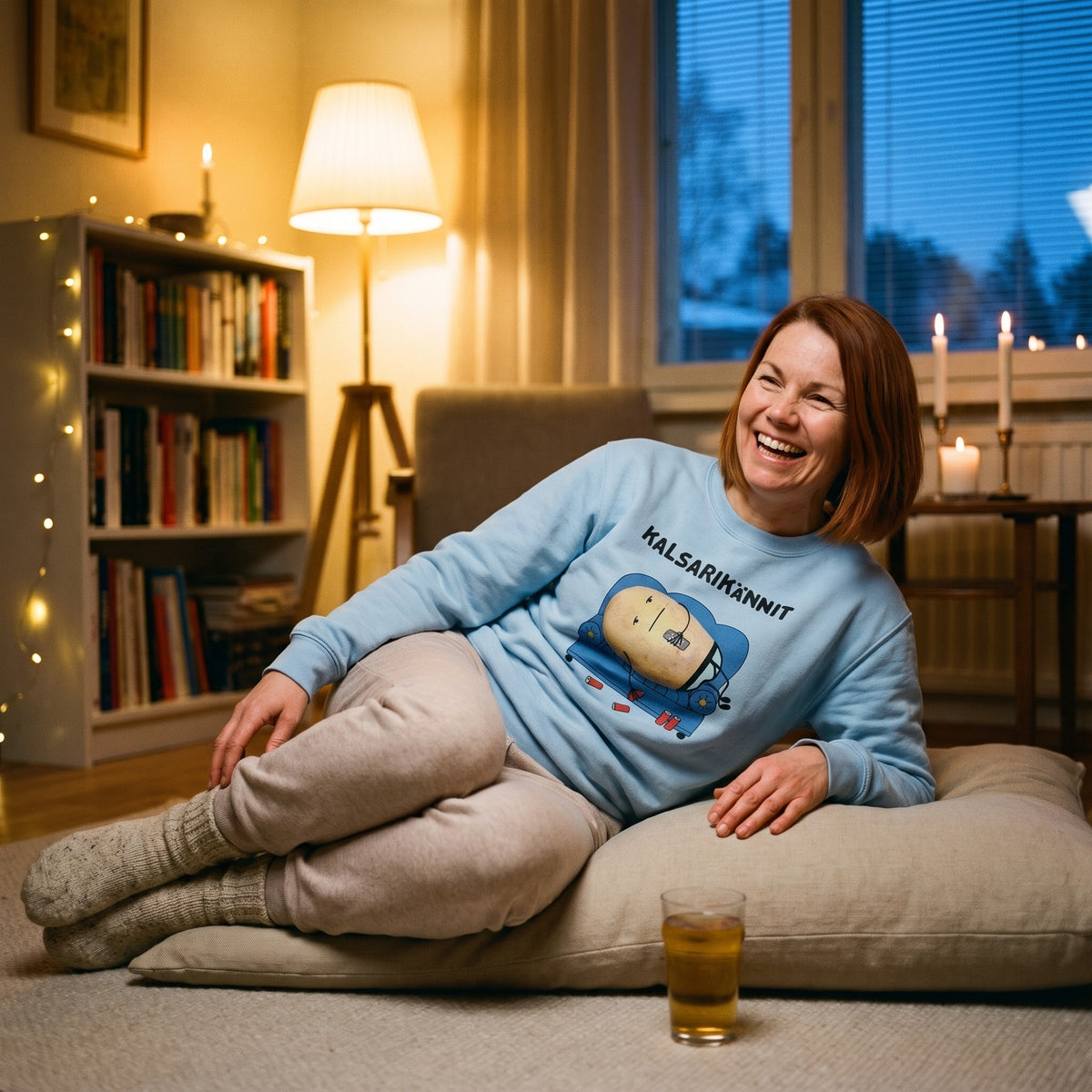 Kalsarikännit sweatshirt in light blue, worn by a laughing woman on a big floor cushion, Very Finnish Problems