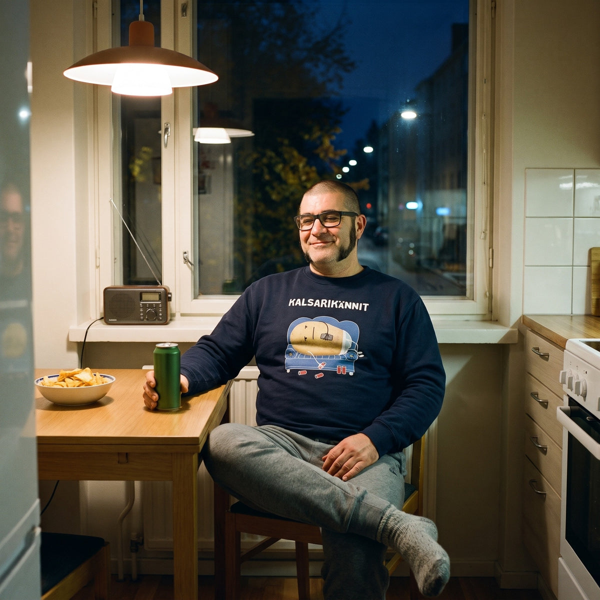 Kalsarikännit sweatshirt in navy, worn by a man in glasses at a kitchen table with crisps, Very Finnish Problems