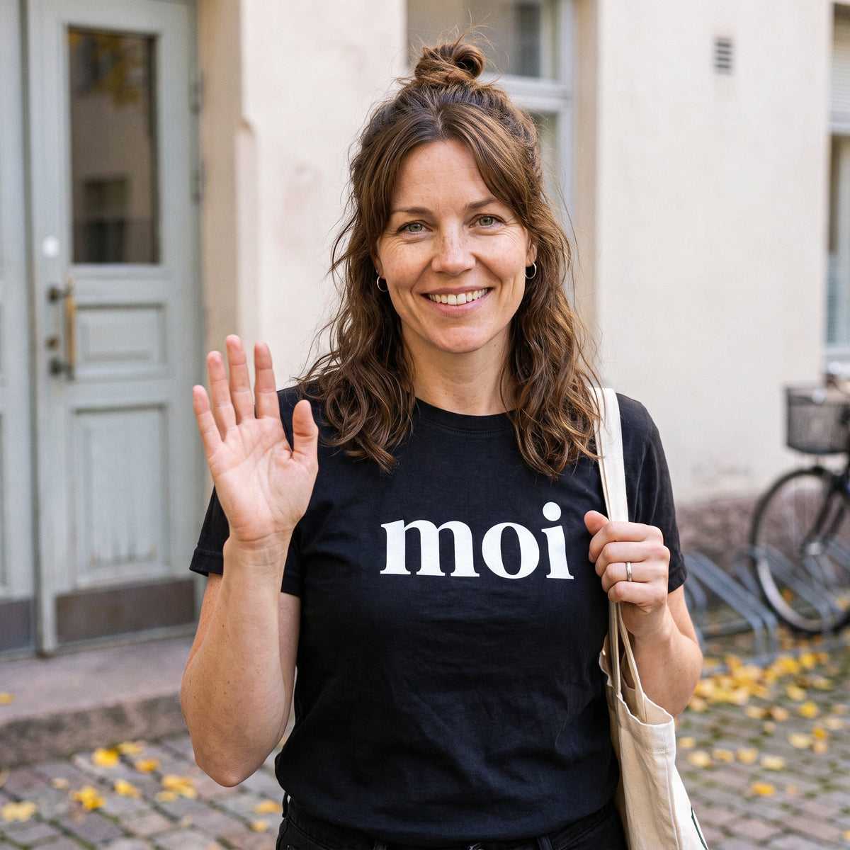 Moi Finnish greeting t-shirt in black, worn by Suvi waving hello to a neighbour in a Helsinki courtyard, Very Finnish Problems