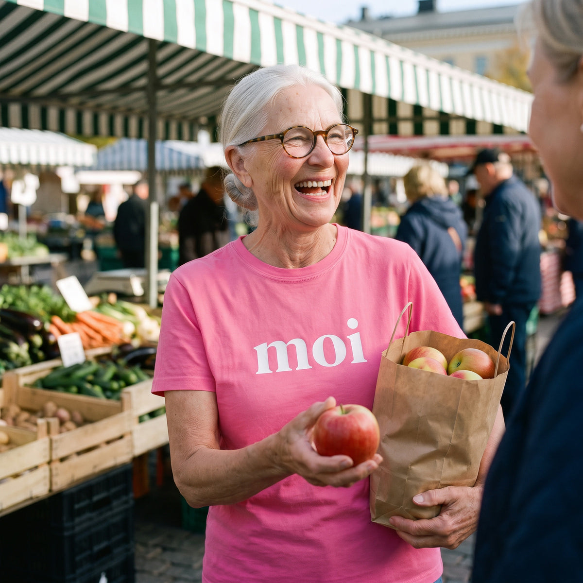 Moi Finnish greeting t-shirt in charity pink, worn by Raija laughing and handing an apple to a friend at a Finnish market, Very Finnish Problems