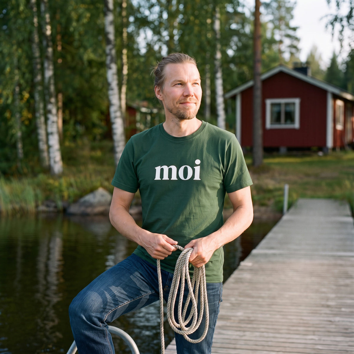 Moi Finnish greeting t-shirt in forest green, worn by Joonas coiling a rope on a lake jetty by a red cottage, Very Finnish Problems