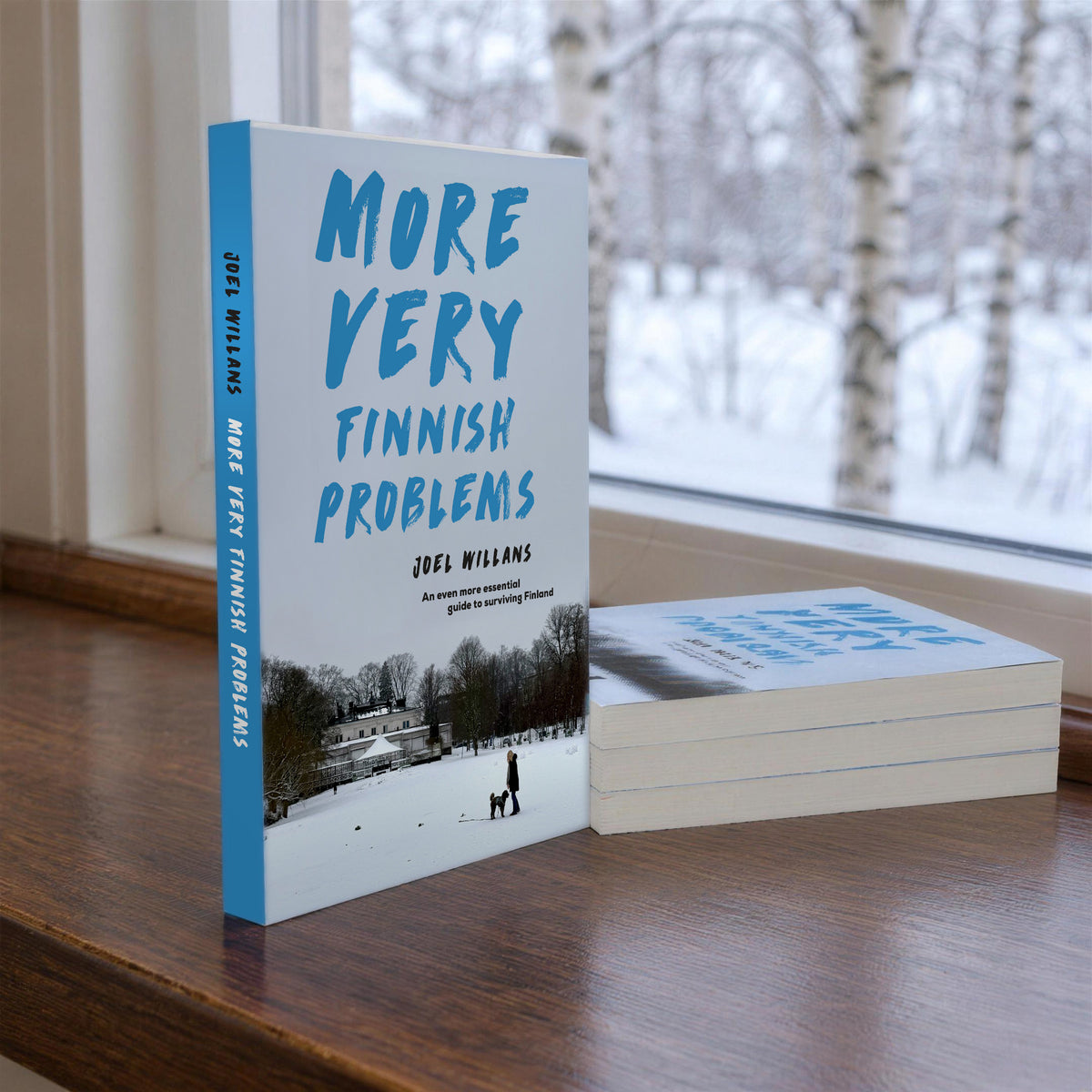More Very Finnish Problems paperback by Joel Willans standing on a windowsill with three more copies stacked beside it, snowy birch trees outside