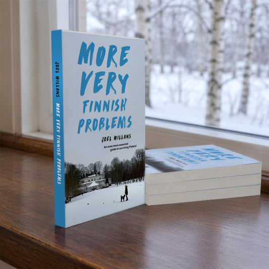 More Very Finnish Problems paperback by Joel Willans standing on a windowsill with three more copies stacked beside it, snowy birch trees outside