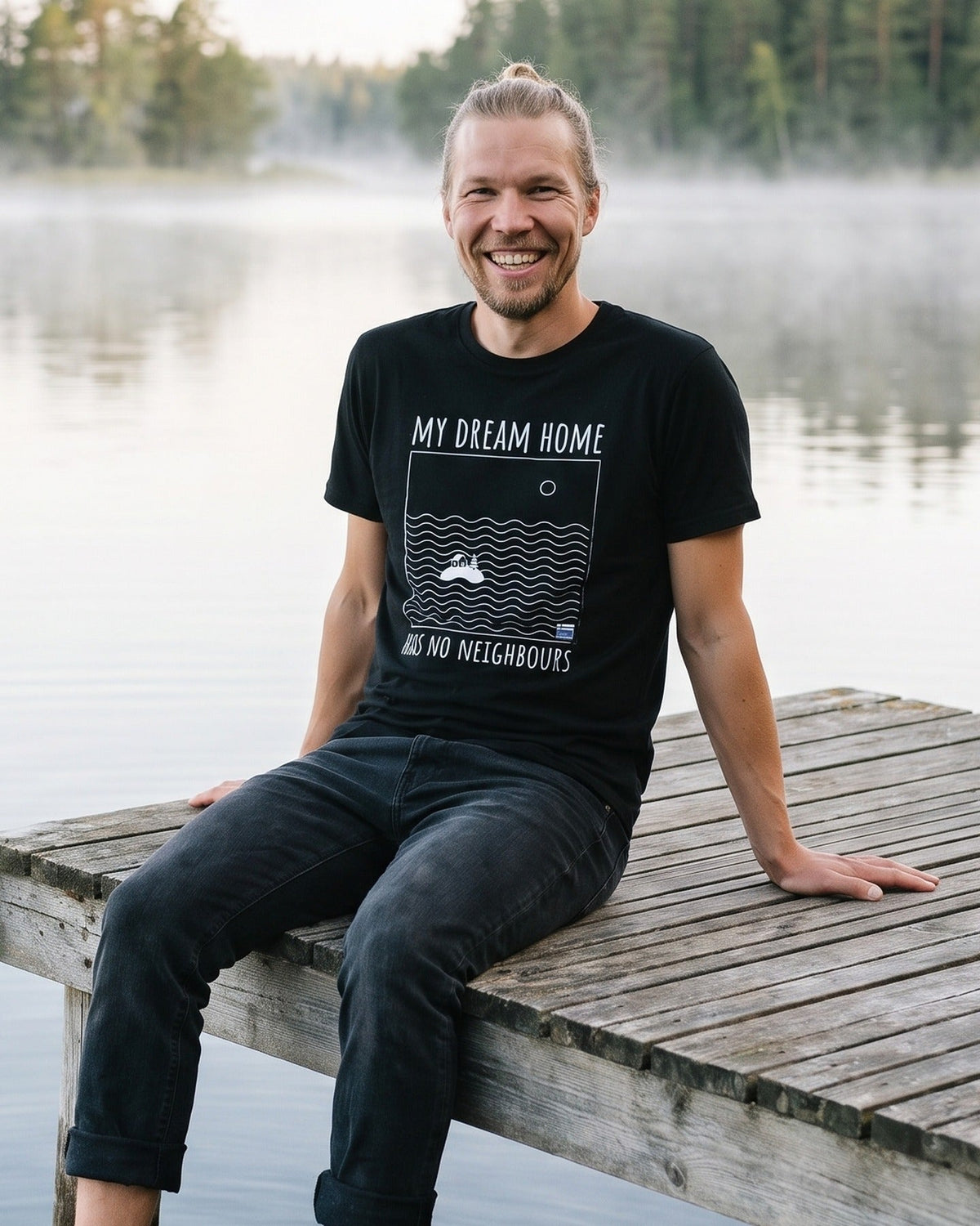 No Neighbours t-shirt in black, worn by Joonas sitting on the end of a lake jetty with his feet in the water, Very Finnish Problems
