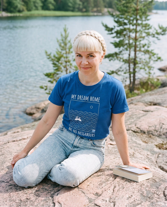 No Neighbours t-shirt in true royal blue, worn by Venla sitting on a granite lake shore with a book, Very Finnish Problems