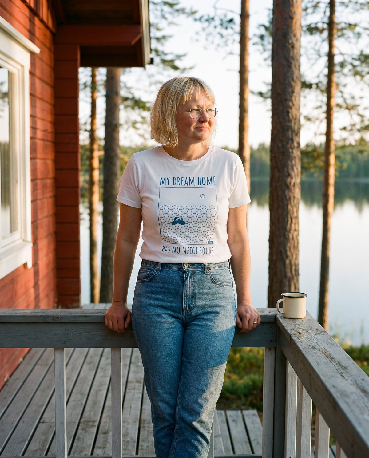 No Neighbours t-shirt in white, worn by Marika leaning on a mökki veranda rail with her morning coffee, Very Finnish Problems