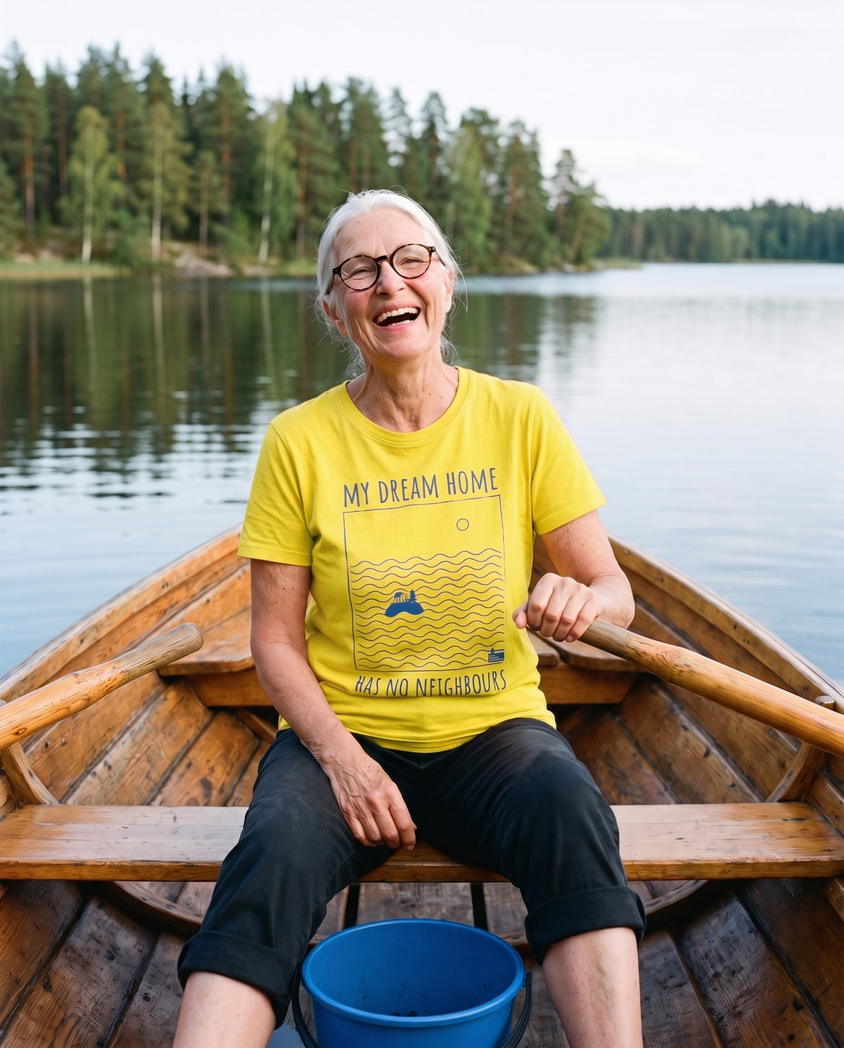 No Neighbours t-shirt in yellow, worn by Raija laughing in a rowing boat on a still Finnish lake, Very Finnish Problems