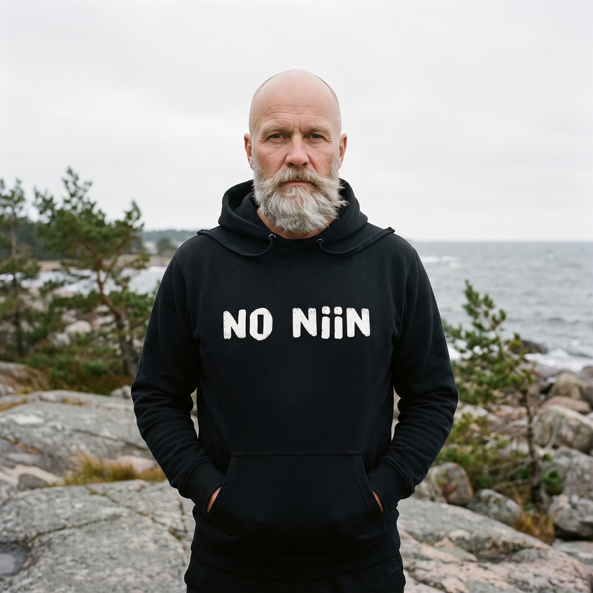 No niin essential eco Finnish language hoodie in black, worn by Mikko on the Finnish Baltic granite shore, Very Finnish Problems