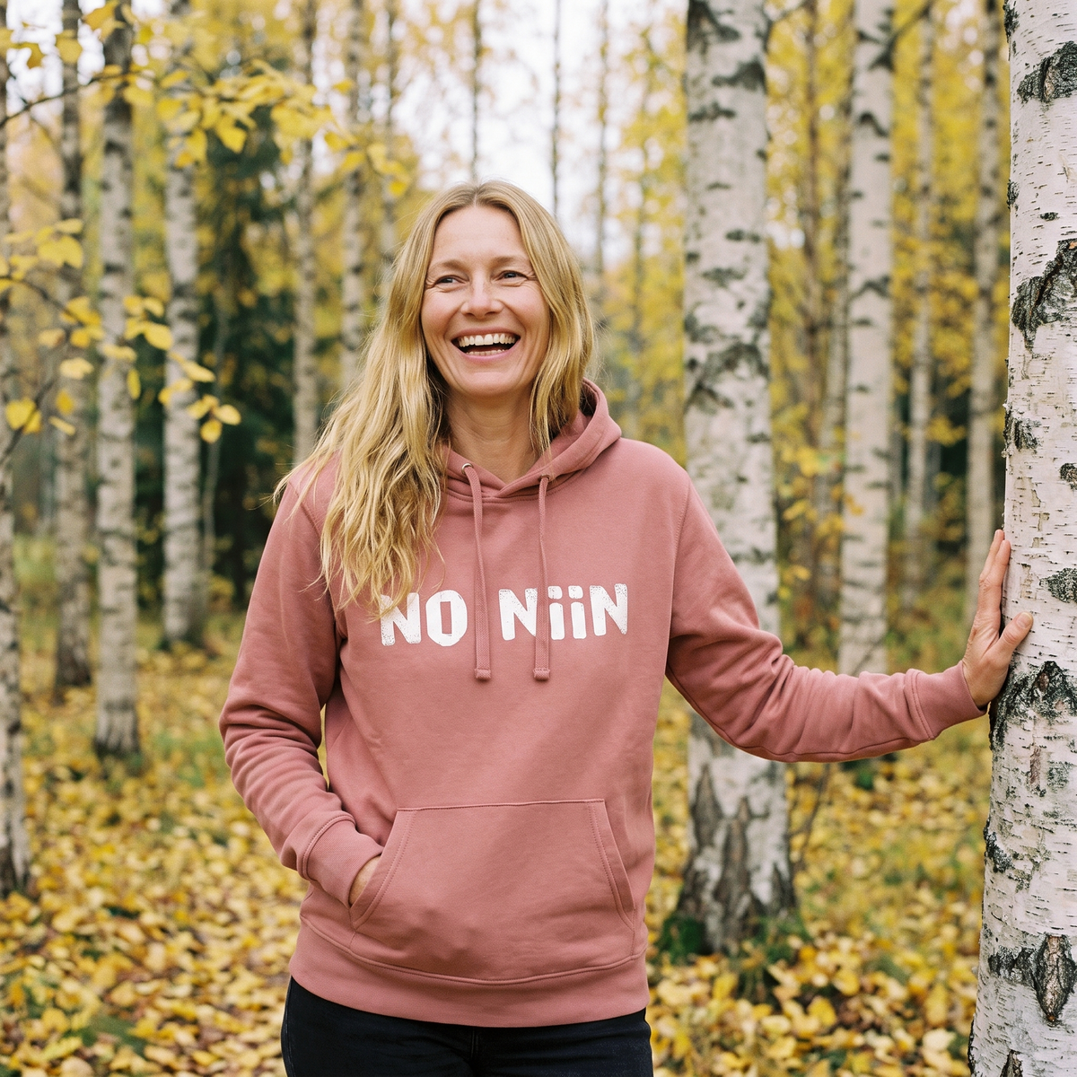 No niin essential eco Finnish language hoodie in canyon pink, worn by Elina laughing in an autumn Finnish birch wood, Very Finnish Problems