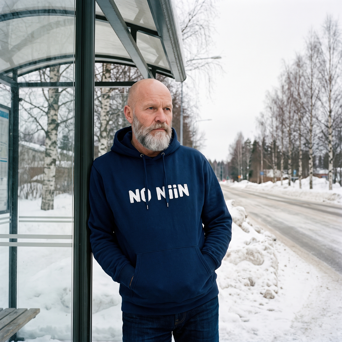 No niin Finnish language hoodie in navy, worn by Mikko waiting at a snowy Finnish bus shelter, Very Finnish Problems