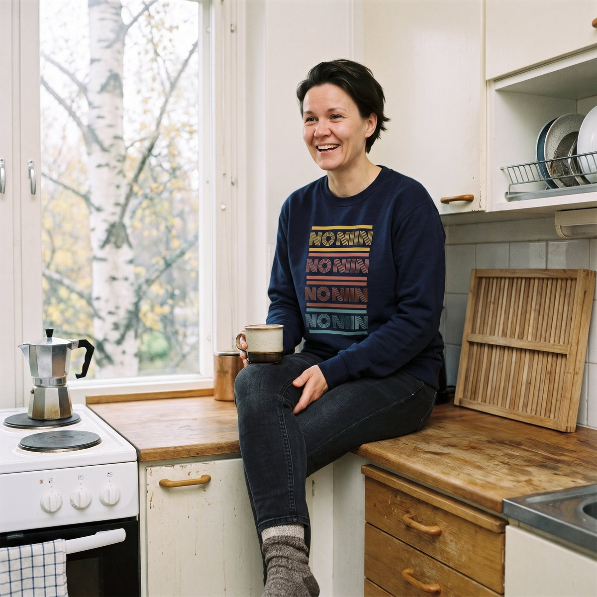 No niin retro sweatshirt in navy, worn by a laughing woman with short dark hair sitting on a Finnish kitchen counter with a coffee mug, Very Finnish Problems