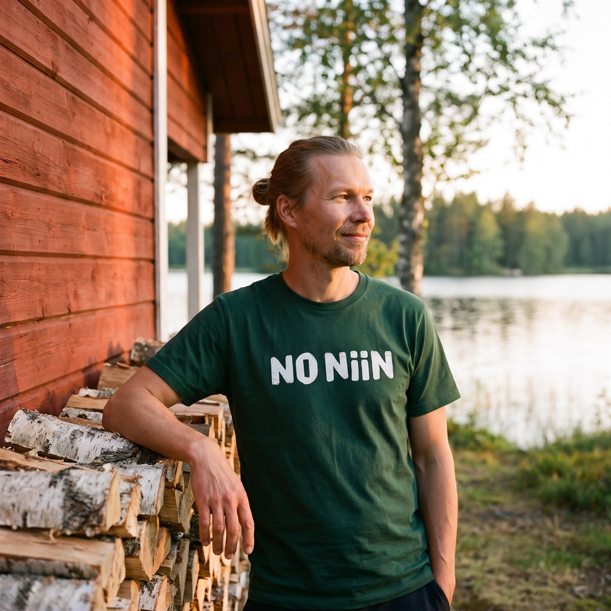 Joonas leaning on a woodpile at a lakeside cottage in the forest green No niin t-shirt, Very Finnish Problems
